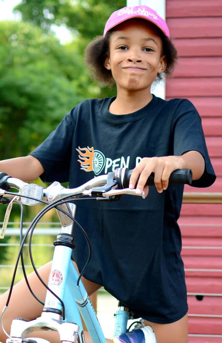 A smiling child on a bike, wearing an Open Roads Bike Program shirt.